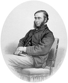 A black and white portrait of William Budd seated on a velvet chair. He is wearing a suit jacket and has a beard but no moustache.