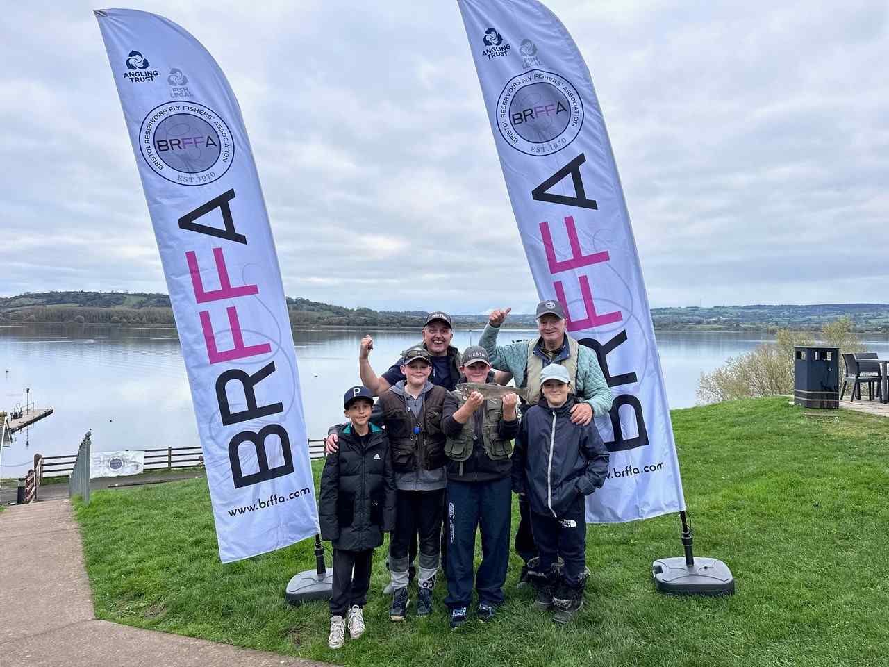 Four children stood with two tutors stood next to a lake holding up a fish they have caught.