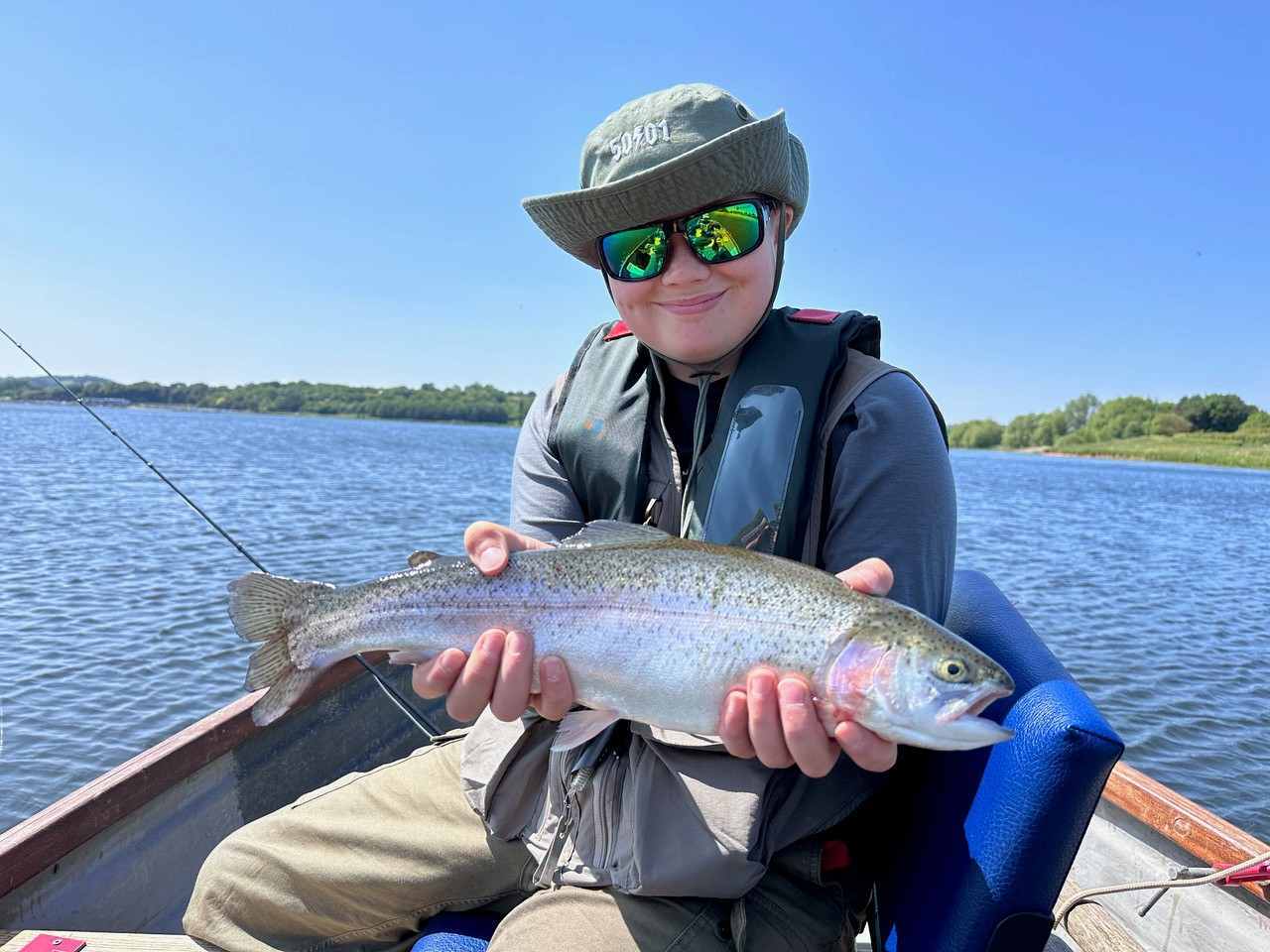 Boy sat on a wooden fishing boat holding up a big fish he has caught.