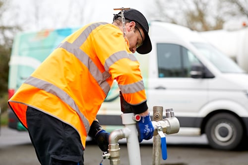A man in a high visibility jacket fitting a water pipe. He is wearing blue gloves, a black bump cap and safety goggles.