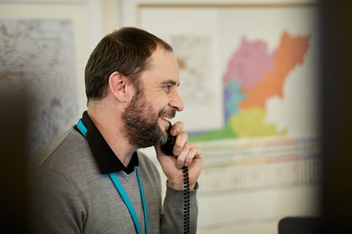 A smiling man with a beard answering the phone. He is wearing a grey jumper and a black polo shirt.