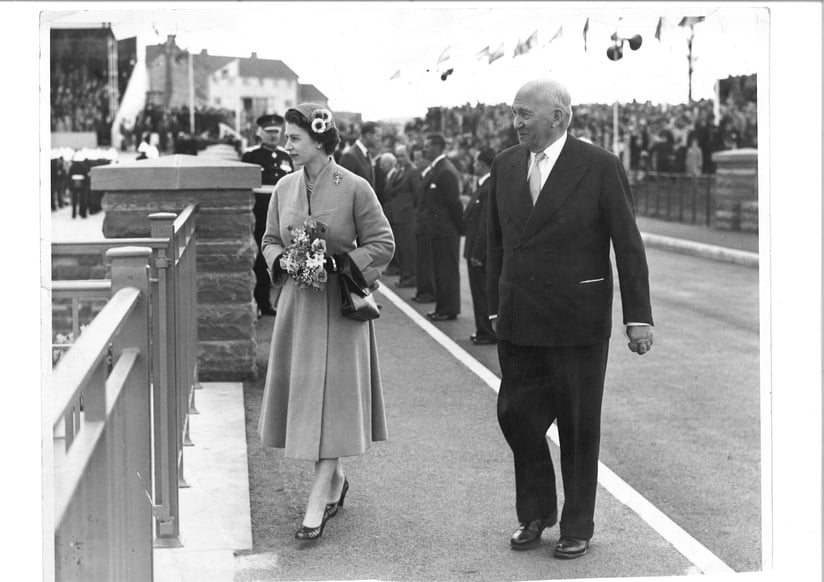 A Black and White photo of Queen Elizabeth the second, wearing a full length coat and a hat. She is holding flowers. She is there to open Chew Valley Lake in front of crowds in April 1956