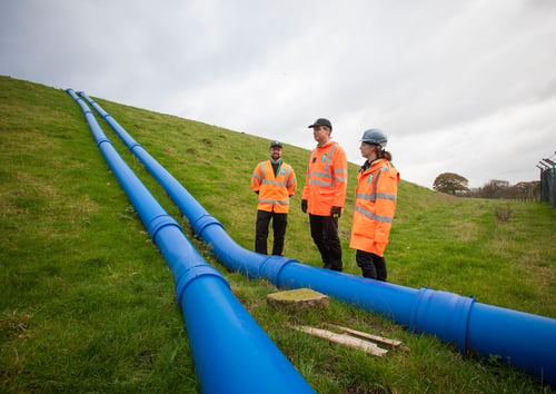 Three Bristol Water employees in high visibility clothing looking at water supply pipe on a grassy hill