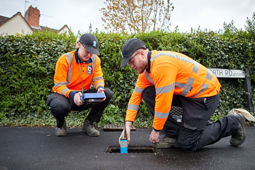 Two Bristol Water employees using remote control equipment to search for leaks. They are wearing high visibility clothing and are crouching by a drain cover.