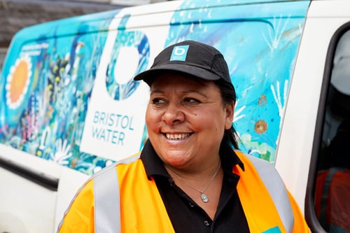 A smiling woman in a Bristol Water bump camp. She is wearing a black polo shirt and an orange high visibility jacket. She is standing in front of a Bristol Water van. 