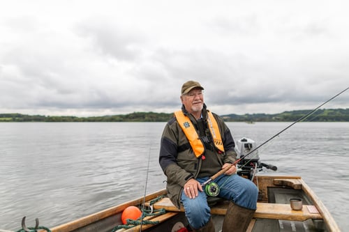 A man with a grey beard and a green hat holding a fishing rod. He is sitting on a boat which is sitting in the middle of a large lake. There are green trees in the background. 
