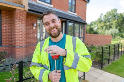 A smiling man with brown hair and a beard, wearing a blue t-shirt and yellow high visibility jacket. He is standing outside of a red brick house