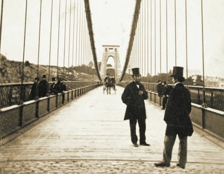 Two men in top hats and suits talking on the finished suspension bridge in a sepia photo. A horse and cart is making its way towards them in the background. A few other suited men are stood on the left side, looking out over the bridge. Two men in top hats and suits talking on the finished suspension bridge in a sepia photo. A horse and cart is making its way towards them in the background. A few other suited men are stood on the left side, looking out over the bridge.