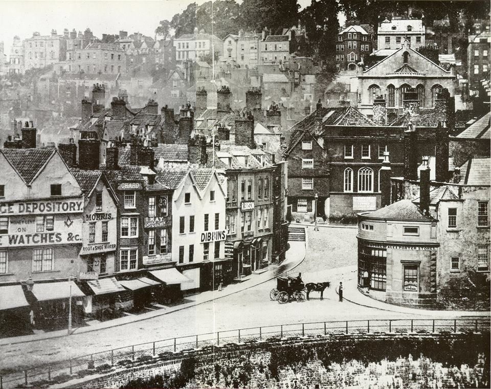 A Black and white image of Bristol in the 1800s showing the bottom of Colston Street and the river Frome before it was blocked off. A horse and cart is in the middle of an empty road, houses and storefronts line the road and the hill in the background.