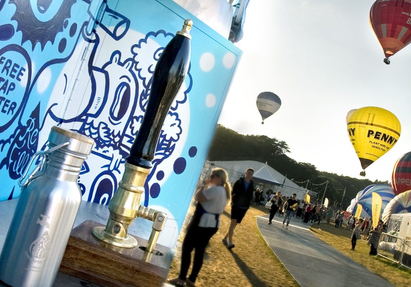 A reusable water bottle next to a water pump at a festival with balloons in the sky behind it
